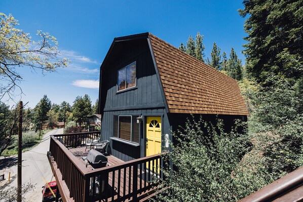 A two-story cabin with a wooden deck, a yellow door, and a brown roof, surrounded by trees.