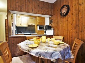 A cozy dining area with a wooden table set for four, surrounded by wooden chairs. A kitchen with wooden cabinets and a microwave is visible in the background.