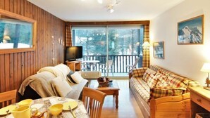 Une pièce chaleureuse avec un décor intérieur en bois, une télévision, un canapé, une table à manger avec de la vaisselle jaune et un balcon offrant une vue sur les arbres.