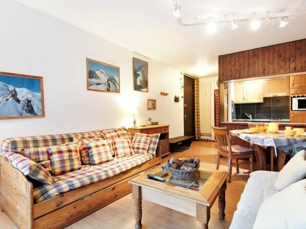 A cozy living room with a plaid sofa, a wooden coffee table, and a kitchen area with wooden cabinets and a dining table.