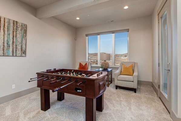 A room with a foosball table, a sofa, a chair, and a window with a view of a building.