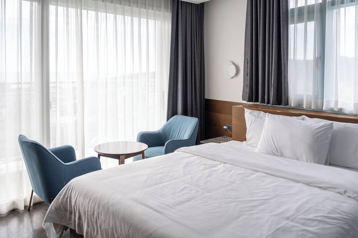 A hotel room with a large bed, a small round table, two blue armchairs, and a view through sheer curtains.