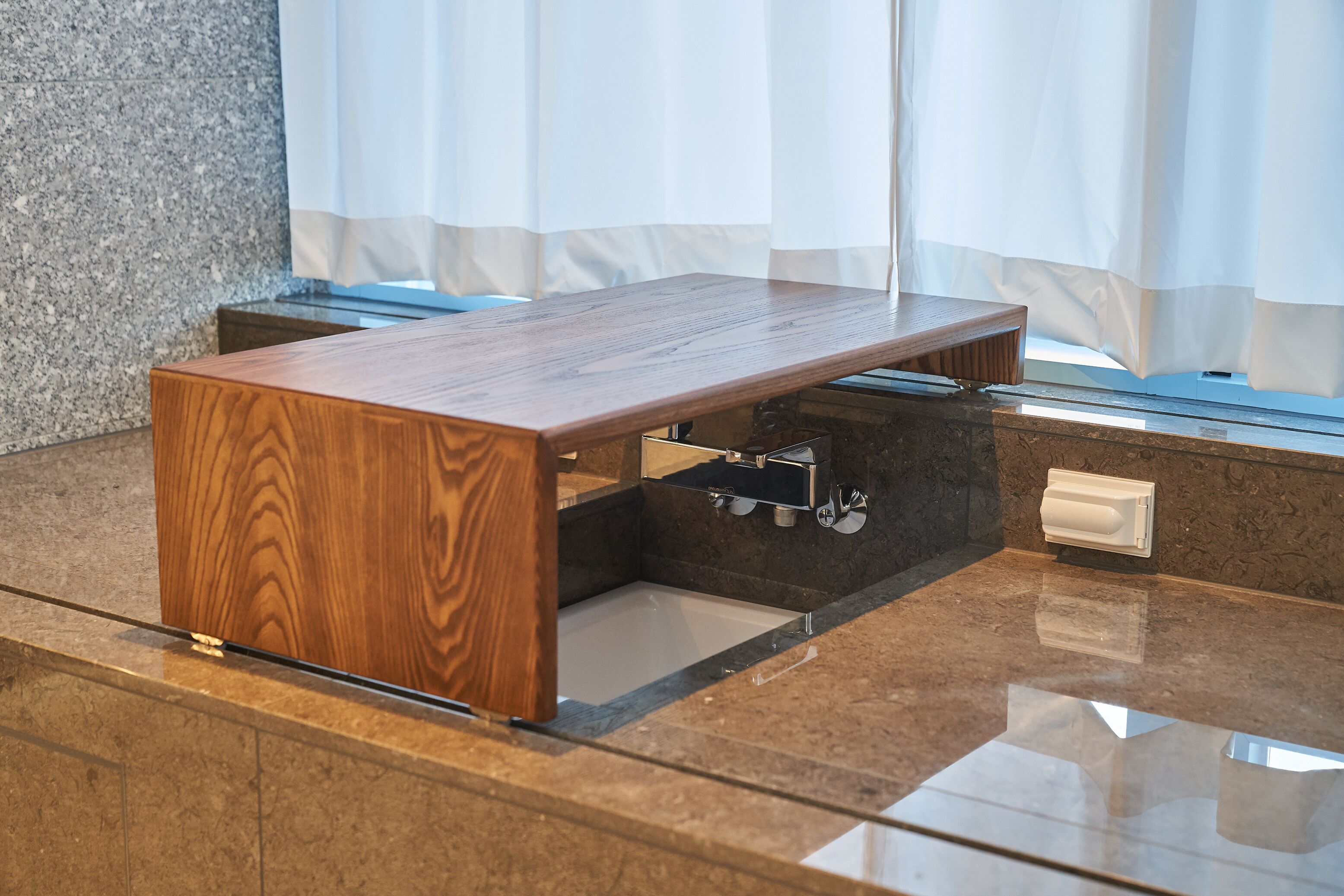 A wooden table with a built-in sink in a modern bathroom with granite countertops and a window with sheer curtains.