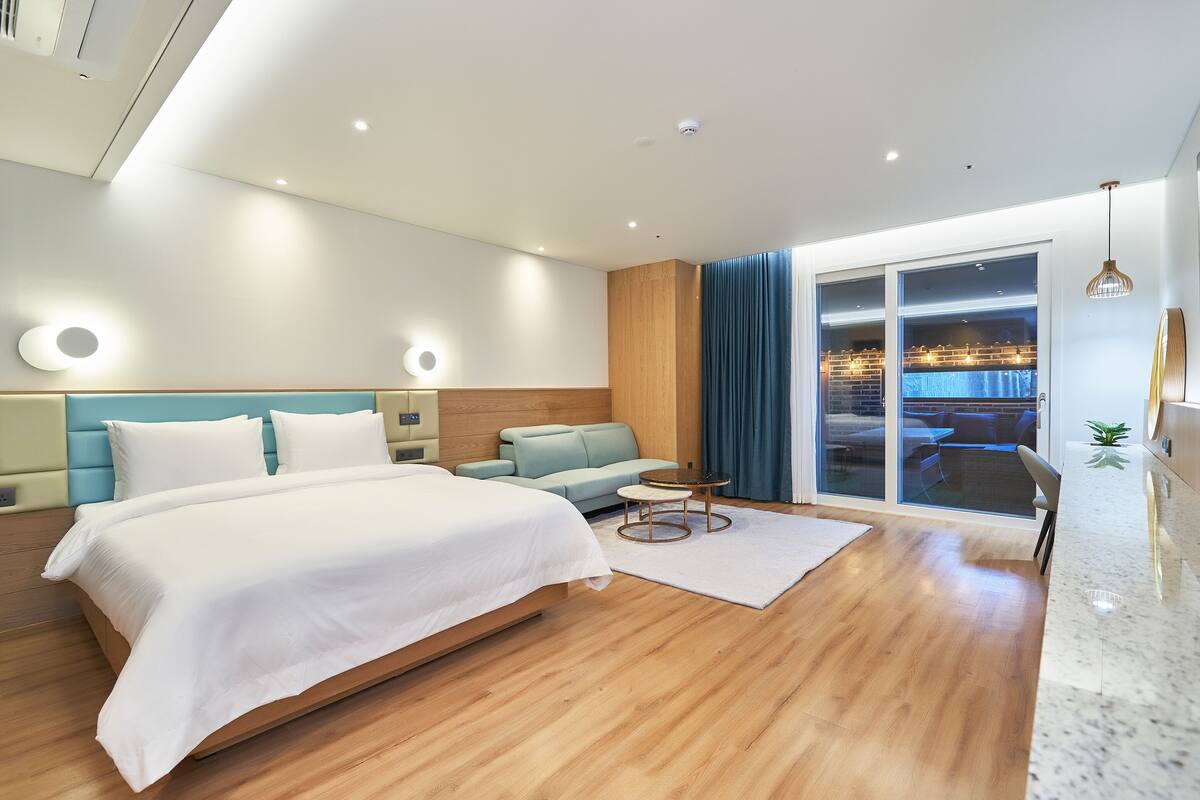 A modern hotel room with a large bed, a sofa, a small table, and a view of the outdoor area through the glass door.