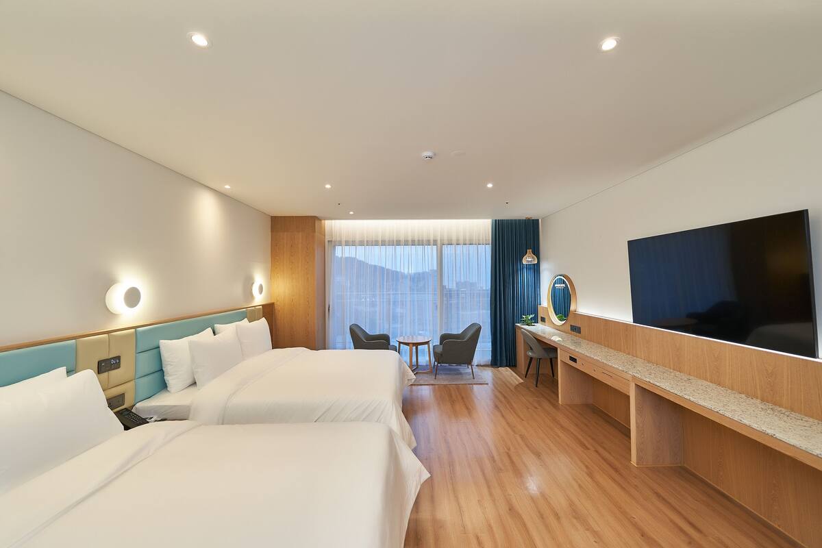 A hotel room with two beds, a desk, a chair, a TV, and a large window with curtains.
