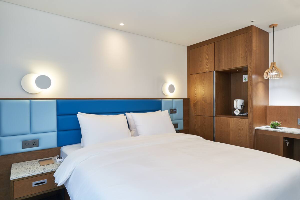 A modern hotel room with a large bed, a wooden wardrobe, a small desk, and a pendant light.