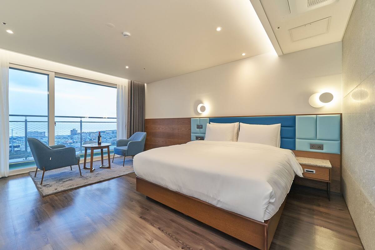 A modern hotel room with a large bed, a seating area with a table, and a city view through large windows.