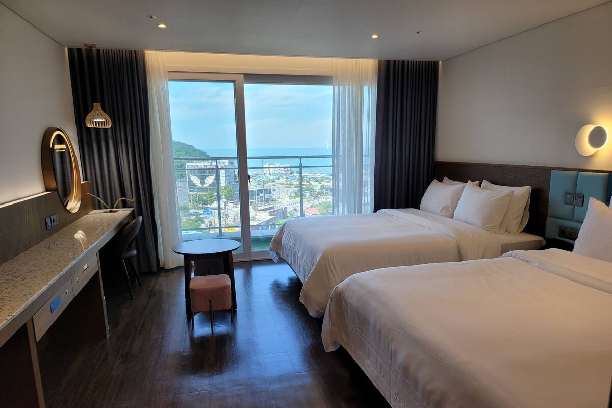 A hotel room with two beds, a desk, a chair, a small table, and a view of the city through large windows.