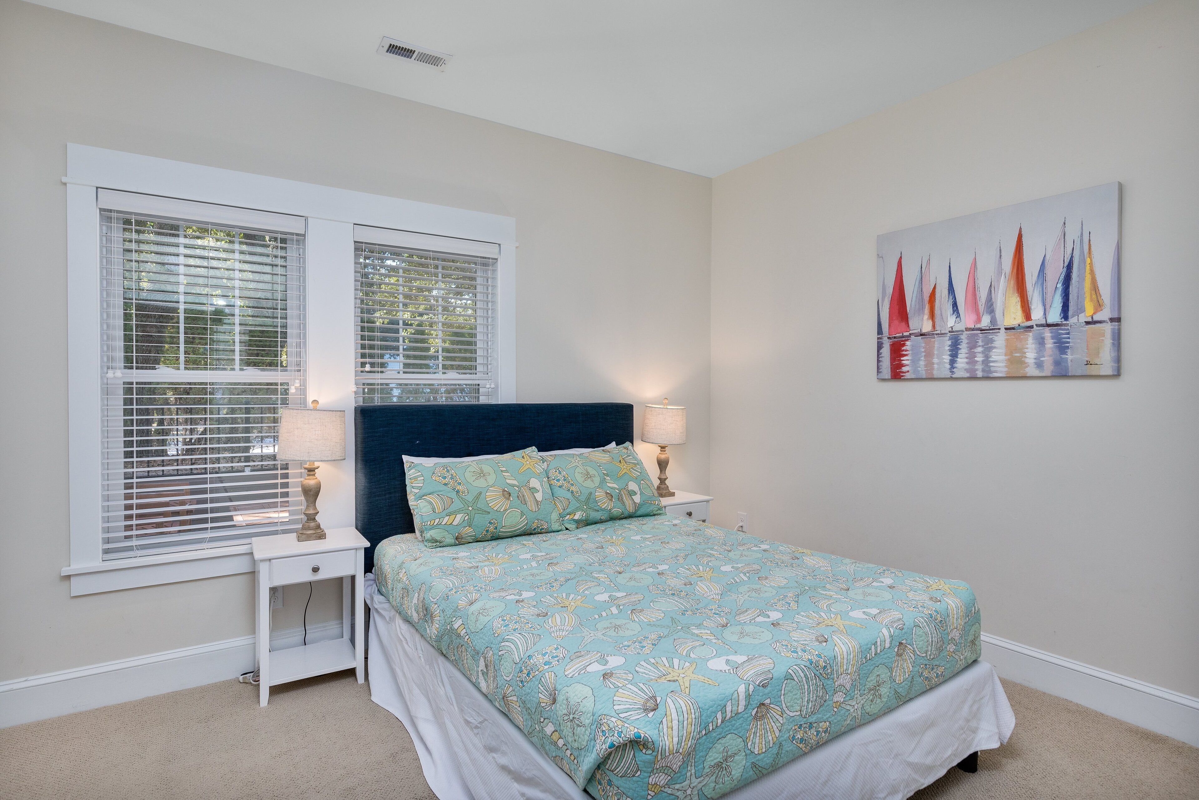 A bedroom with a bed, two bedside tables, a nightstand, a window with blinds, and a wall-mounted painting.