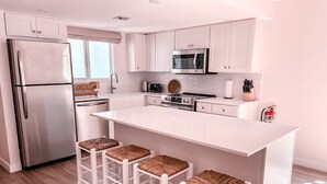 A modern kitchen with white cabinets, stainless steel appliances, a large island with bar stools, and a wooden floor. - Luxury condo steps from one of the world's best beaches. (George Town)