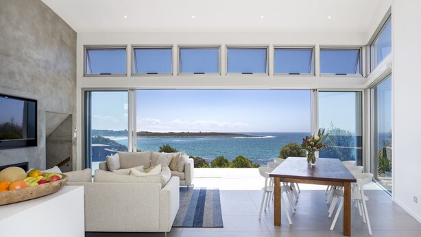 Una sala de estar moderna con un gran ventanal con vistas al mar, un sofá, una mesa de comedor y un frutero.