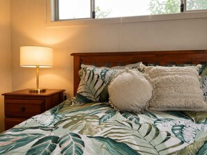 A bed with a leaf-patterned bedspread, a bedside lamp, and a window with a view of trees.