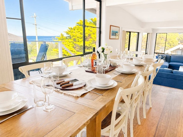 A dining area with a long table set for a meal, large windows with a view, and a blue sofa in the background. - Scotts Ocean View at Mollymook (Mollymook)