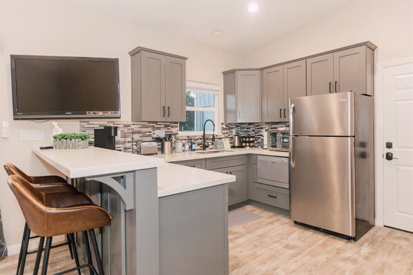 A modern kitchen with grey cabinets, a stainless steel refrigerator, a flat-screen TV, and a bar stool.