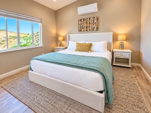 A bedroom with a large bed, two bedside tables, a wall-mounted air conditioner, and a window with a view of a landscape.