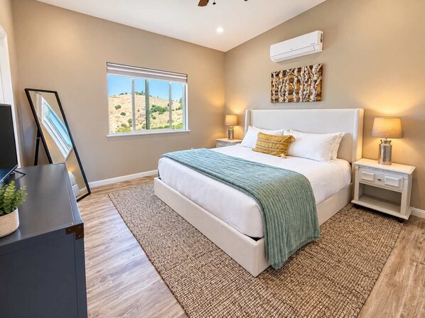 A bedroom with a bed, bedside lamps, a window with a view, and an air conditioning unit.