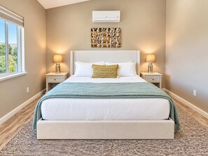 A bedroom with a large bed, two bedside tables, a wall-mounted air conditioner, and a window with a view.