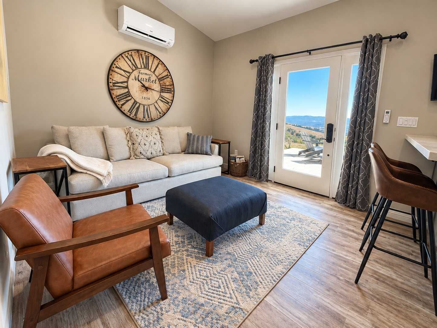 Un salon comprenant un canapé, une table basse, un fauteuil en cuir, une horloge et une vue sur l’extérieur, visible à travers une porte vitrée.