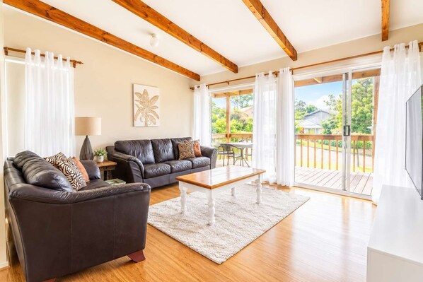 A living room with a leather sofa, a coffee table, and a TV. There is a wooden deck outside the room.