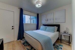 A bedroom with a bed, a nightstand, a window with curtains, and a wall with framed pictures.