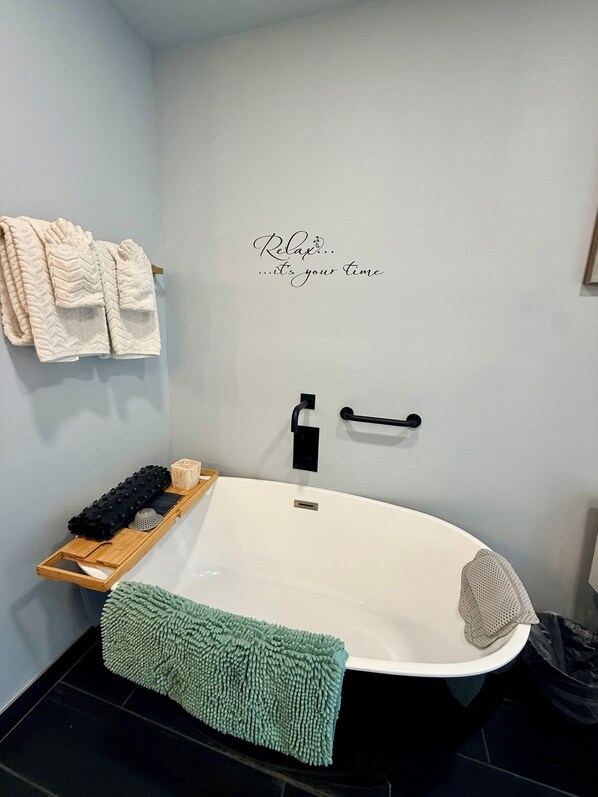 A modern bathroom with a freestanding bathtub, a towel rack with folded towels, a wooden tray with bath products, and a green bath mat.