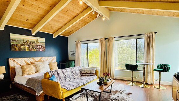 Habitación moderna con cama, sofá, mesa de centro y vistas a zonas verdes a través de amplios ventanales.