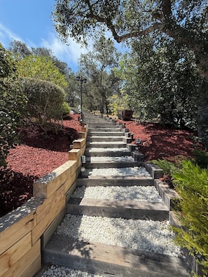 A stone staircase with wooden railings, surrounded by red mulch and white pebbles, leading up to a shaded area with trees.