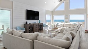 A spacious living room with a large sectional sofa, a flat-screen TV mounted on the wall, and a view of the ocean through large windows. - Modern Beach Front Home with Volleyball net | Summer's Point by Kaiser Vacation Rentals. (Gulf Shores)