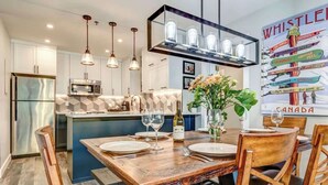 Una cocina moderna con una isla central, luces colgantes y un comedor con mesa y sillas de madera.