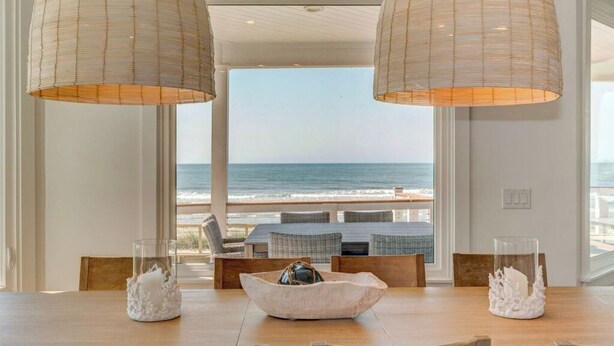 Uma área de refeições com vista para o oceano, com uma mesa, cadeiras, duas luminárias pendentes e vasos decorativos.