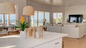 Uma sala de estar ampla com uma televisão grande, sofás brancos e vista para o mar.
