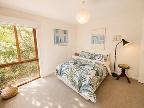 Un dormitorio con un ventanal grande, una cama con colcha de estampado tropical, una mesita de noche, una lámpara de pie y decoración en la pared.