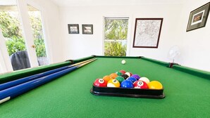 Una mesa de billar con un juego de bolas, una vara de billar y un ventilador en una habitación con ventanas y cuadros en las paredes.