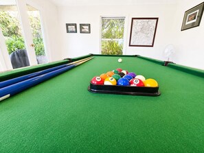 Une table de billard avec un jeu de billes, une queue de billard et un ventilateur, dans une pièce dotée de fenêtres et dont les murs sont ornés de tableaux encadrés.