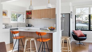 A modern kitchen with a central island, bar stools, a refrigerator, and a window with a view of the outside. - Retro Beach House at Mollymook (Mollymook)