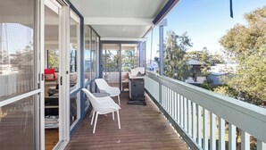 Une véranda couverte avec une terrasse en bois, des chaises blanches, un barbecue et une vue sur le quartier.