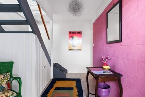 Un pasillo con una escalera, un cartel en la pared, una mesita con un florero y una silla con un cojín colorido.