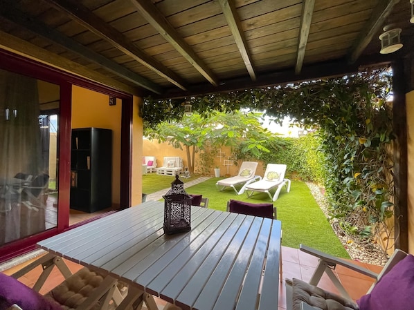 A patio with a table and chairs, a lantern, and a view of a garden with lounge chairs and a lawn.