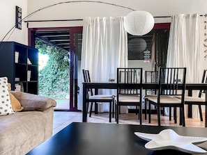 A modern living room with a sofa, a coffee table, and a dining area with a table and chairs. There is a bookshelf and a large window with curtains.