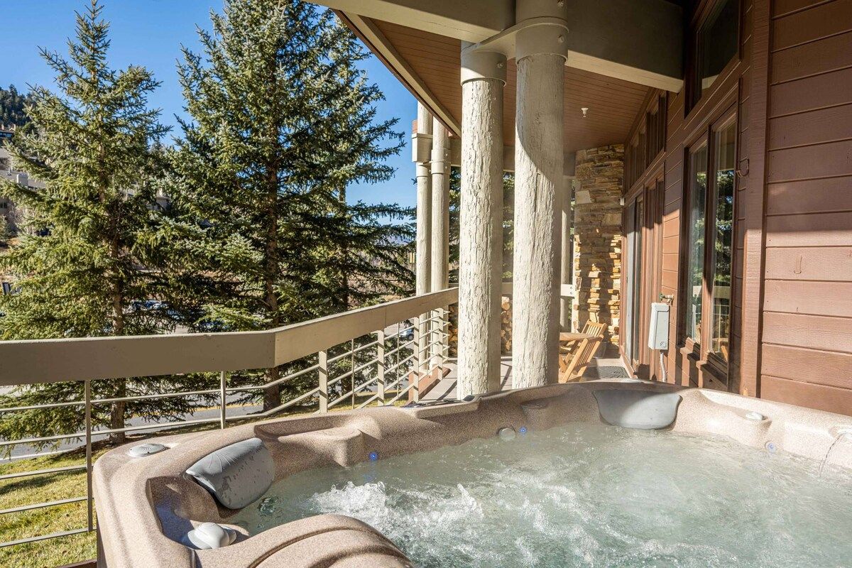 A hot tub area with a stone wall backdrop, a wooden deck, and a view of trees.