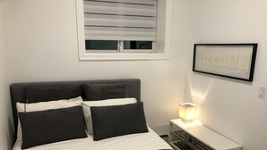 A bedroom with a bed, pillows, a bedside table with a lamp, a window with blinds, and a framed picture on the wall. - Amazing 1 Bedroom suite in Vancouver (Vancouver)