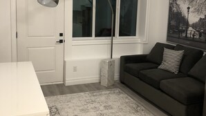 A modern living room with a white door, a grey sofa, a black and white patterned pillow, a grey and white rug, and a large window with blinds. - Amazing 1 Bedroom suite in Vancouver (Vancouver)