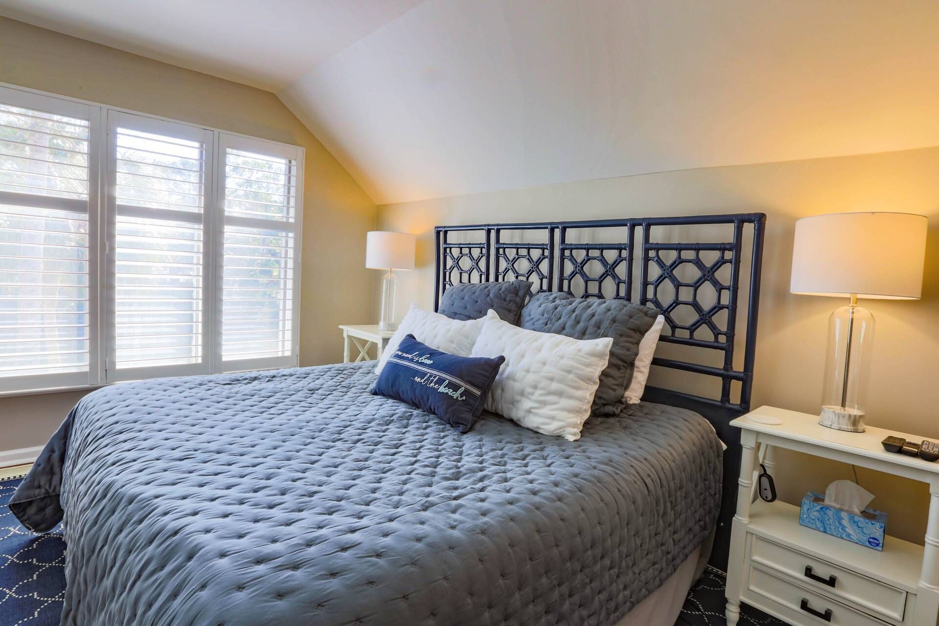 A neatly made bed with a grey quilt, a headboard with a geometric pattern, bedside lamps, and a small bedside table with a tissue box.