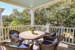 A covered outdoor patio with wicker furniture and a round table, surrounded by lush greenery and trees.