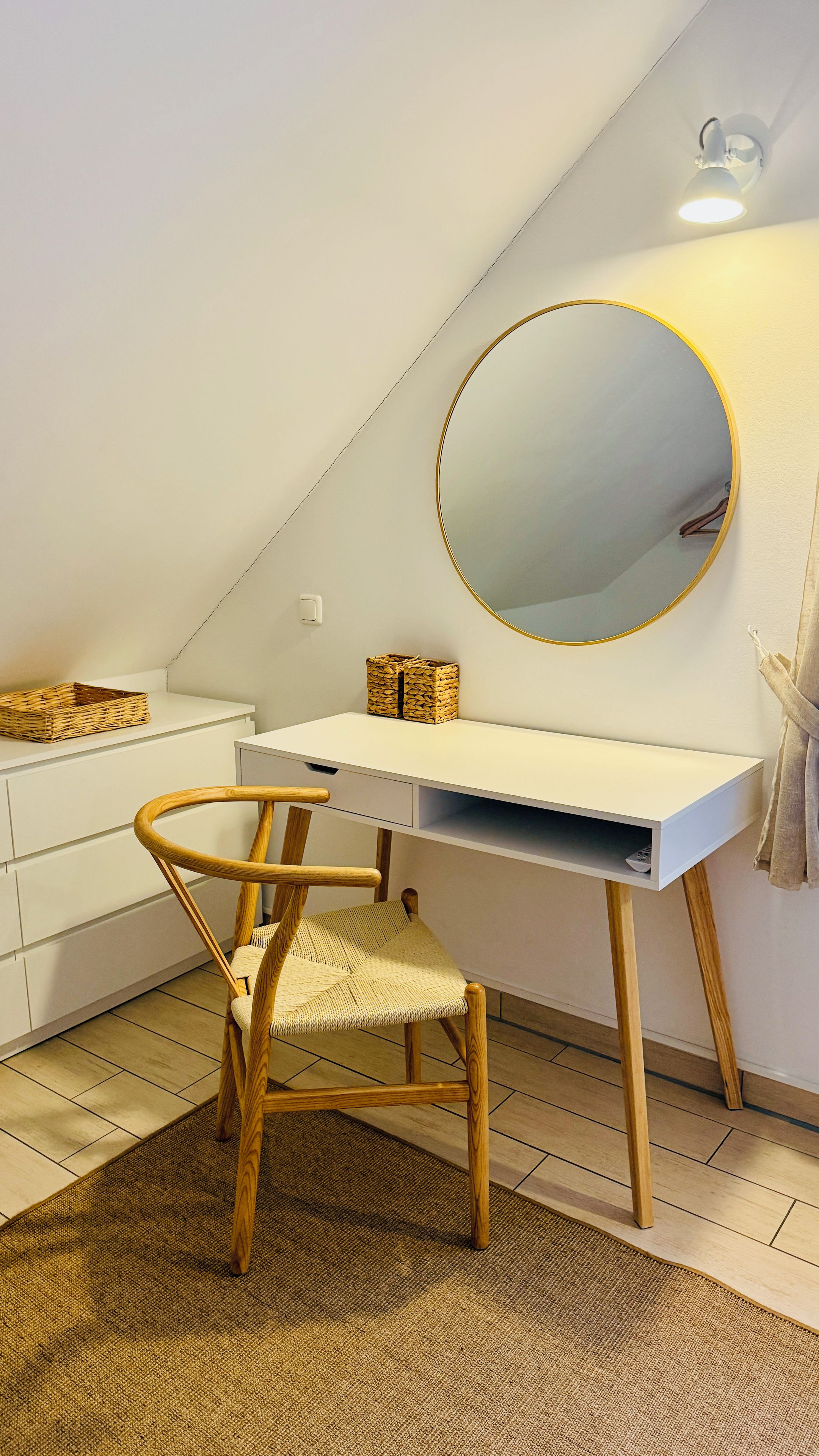 Ein Zimmer mit einem weißen Schreibtisch, einem runden Spiegel, einem gewobenen Stuhl und einem kleinen Korb auf dem Schreibtisch.