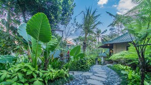 A stone pathway leading through a lush garden with large tropical plants and a traditional house with a tiled roof. - D'Junas Villas by GenuineHost  (Ubud)