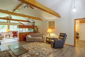 A cozy living room with a sofa, a chair, a coffee table, and a rug. There is a window with curtains, a ceiling fan, and a wall-mounted lamp. - Nature Lover's Cottage on Wooded Acreage (Nova)