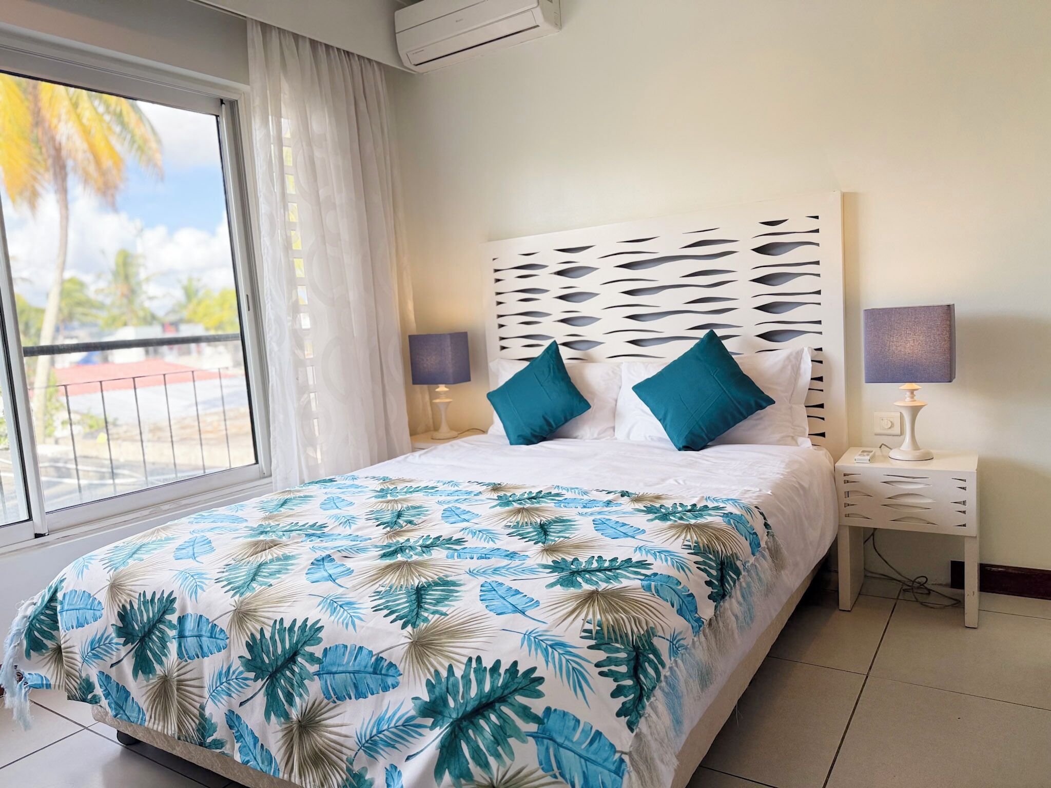 A hotel room with a bed, two bedside lamps, a window with curtains, and a view of palm trees.