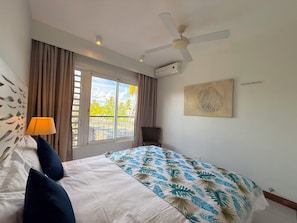A bedroom with a bed, a ceiling fan, a window with curtains, and a wall-mounted air conditioner. - PLAGE BLEUE BEACHFRONT APARTMENTS BY LOV (Pointe Aux Piments)