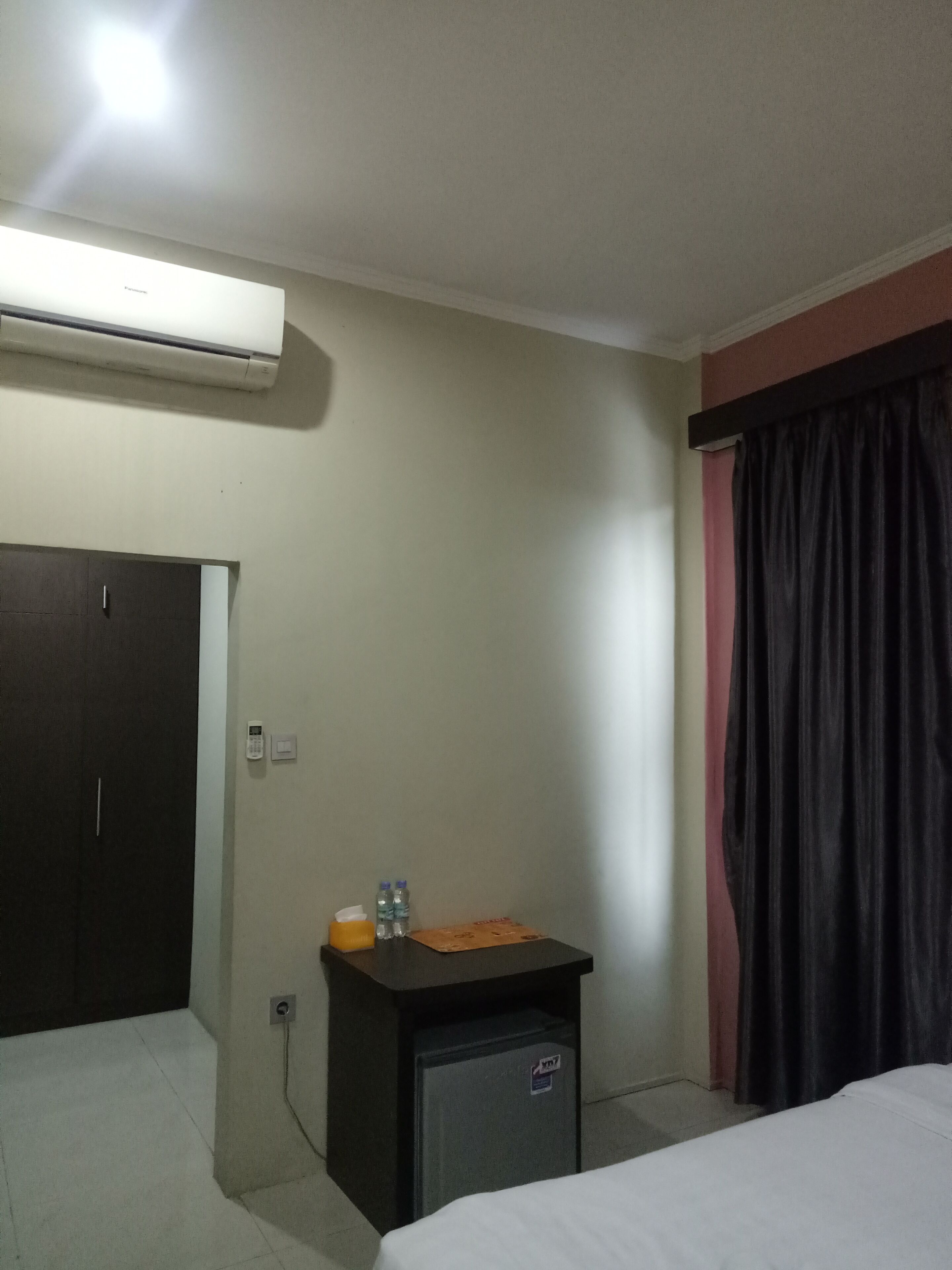 A hotel room with a bed, a small desk with a telephone and water bottles, an air conditioning unit, and a door.